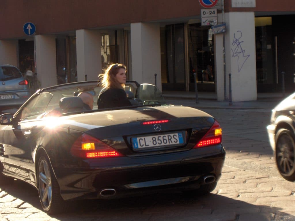 Eine Frau sitzt in einem Auto auf einer Straße in der Nacht, mit Gebäuden, einer Säule, einem Schild und anderen Fahrzeugen im Hintergrund.