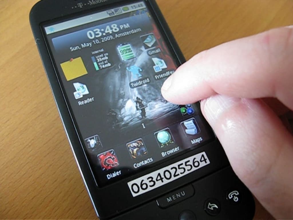 Eine Person nutzt ein Mobiltelefon auf einem Tisch, mit mehreren Apps auf dem Bildschirm und einem Zettel mit einer Nummer, der an das Telefon geheftet ist.