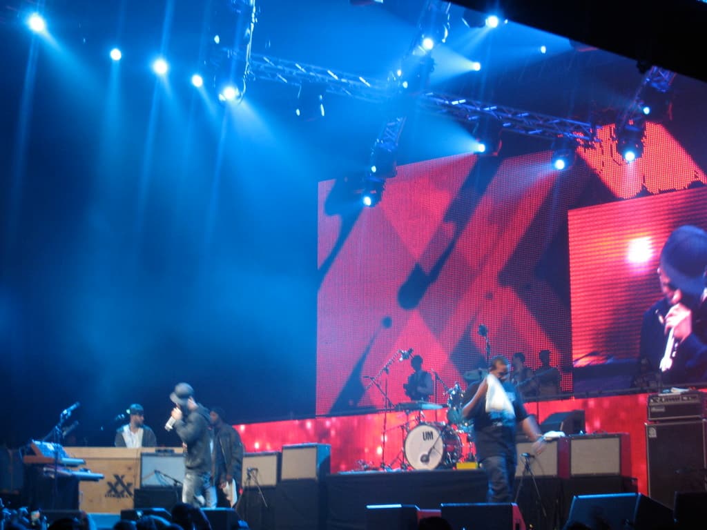 Eine Gruppe von Menschen, die auf einer B├╝hne Musikinstrumente spielen, mit einem Bildschirm dahinter und Lichtern an der Decke.