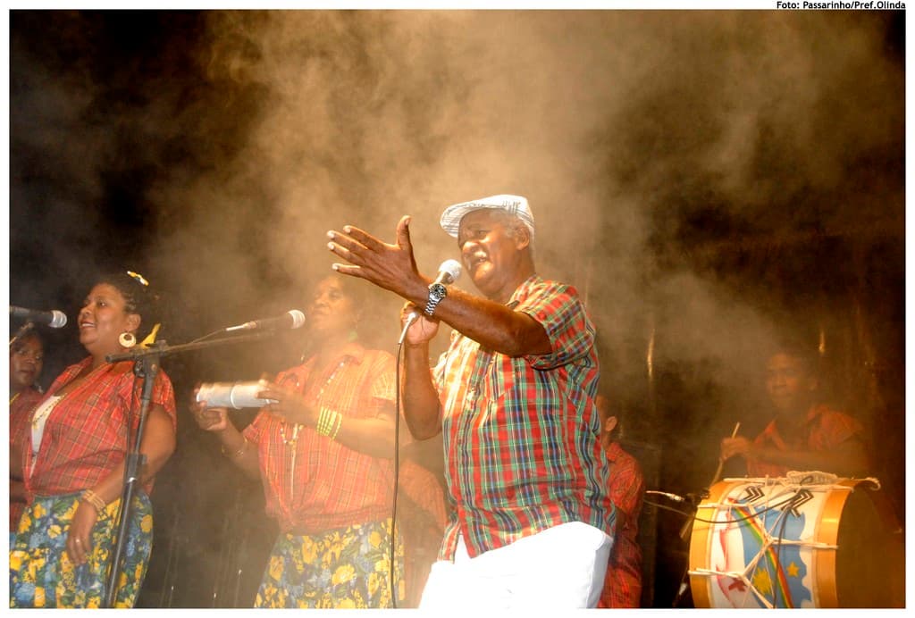 Eine Gruppe von Musikern, die auf der B├╝hne stehen, wobei ein Mann in der Mitte in ein Mikrofon singt und ein anderer Mann rechts ein Schlagzeug spielt.