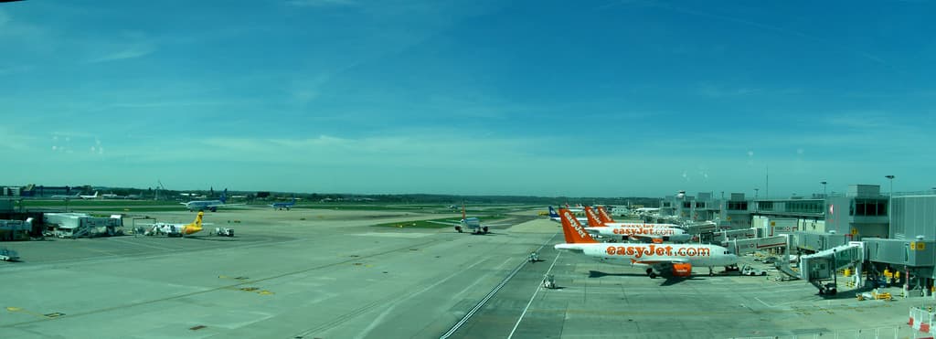 Eine Flughafen-Szene mit Flugzeugen, Gebäuden, Masten, Fahrzeugen und Bäumen unter einem Himmel mit einer Straße unten und einem Zaun im Vordergrund.