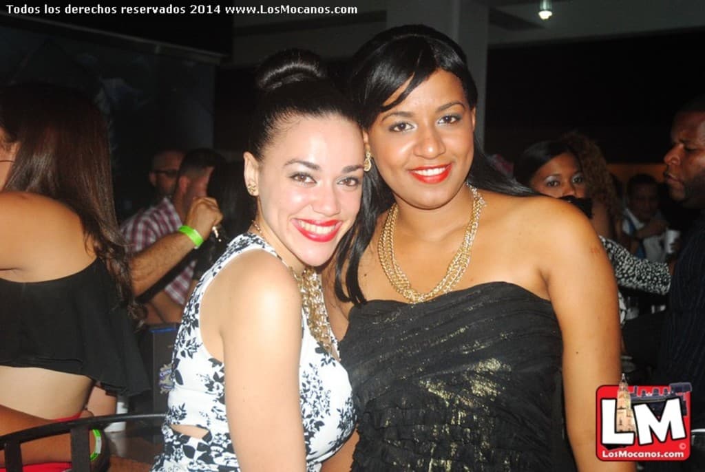 Zwei Frauen in modischer Kleidung lächeln zusammen in einer Bar, mit anderen tanzenden Menschen im Hintergrund und Lichtern an der Decke.