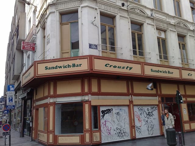 Ein Gebäude mit Wänden, Fenstern, Glastüren, Geländern, Namensschildern und Geschäften, mit einem Pfahl auf der rechten Seite, einem Müllcontainer und Pfählen mit Schildern auf der linken Seite.