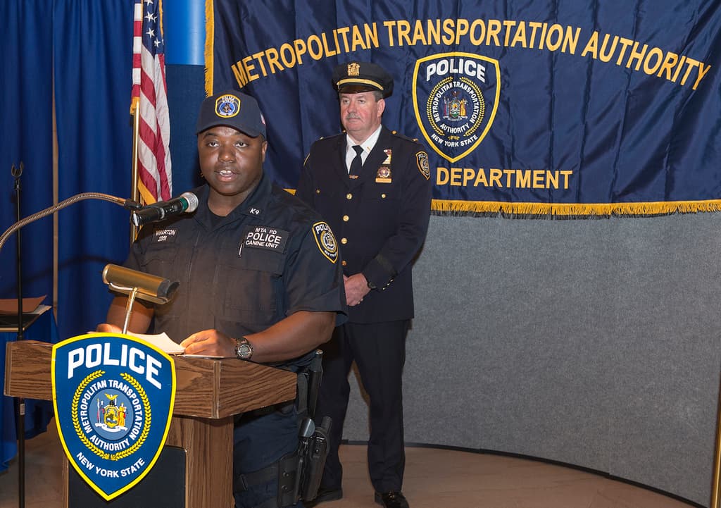 Zwei Polizeibeamte stehen vor einem Rednerpult, einer h├Ąlt Papiere und spricht in ein Mikrofon, mit einer Fahne, einem Tisch und einer Tafel mit Symbolen und Text im Hintergrund.