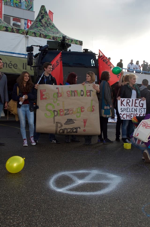 Eine Stadtstraßenszene mit einem Ballon im Vordergrund, Menschen mit Schildern und Fahnen in der Mitte, einem Gebäude und einem Zelt im Hintergrund, unter einem bewölkten Himmel.