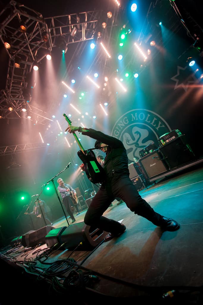 Ein musikalisches Konzert mit drei Personen auf der Bühne: zwei Gitarren spielende Personen rechts mit Mikrofonen davor, ein Hintergrund mit 'MOLLEY' geschrieben und zwei Schlangenlogos, sowie Bühnenlichter oben.
