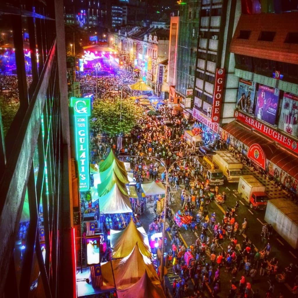 Eine belebte Karnevals-Szene mit einer großen Menge auf der Straße, umgeben von Zelten, Geschäften, Gebäuden und Bannern, mit zahlreichen Lichtern im Hintergrund.