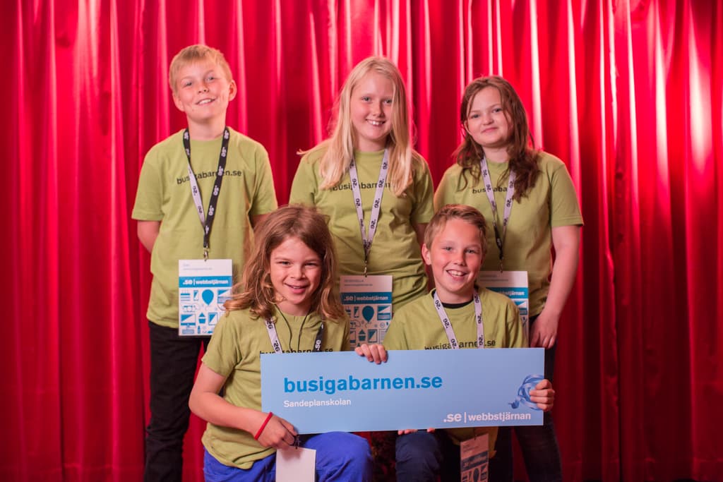Eine Gruppe von Kindern mit ID-Karten, wobei ein Junge und ein Mädchen ein Schild mit Text halten, vor einem Vorhang.
