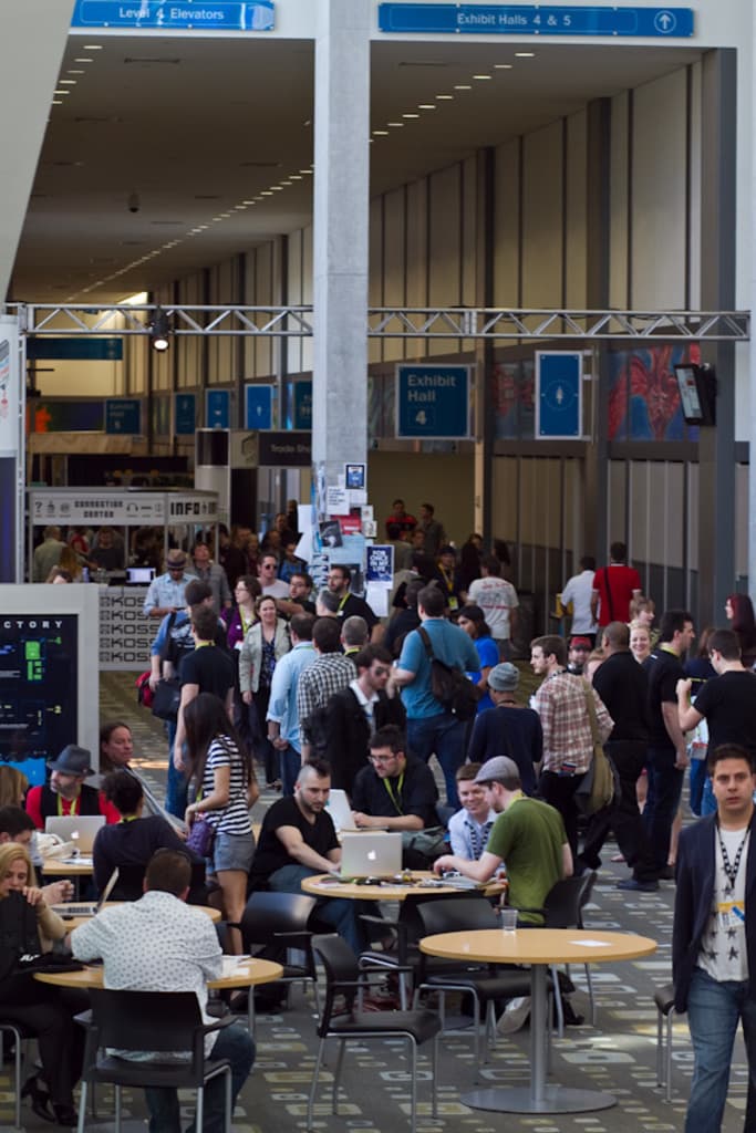 Ein belebter Airport-Lobby mit wartenden Menschen, einige stehen und andere arbeiten, Richtungsschilder an der rechten Wand und Decke sowie Deckenleuchten.