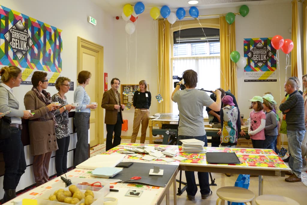 Ein voller Raum mit feiernden Menschen, Tischen mit verschiedenen Gegenständen wie Büchern und Kartoffeln, ein Mann mit einer Kamera auf einem Stativ, Menschen mit Tassen in der Hand und Kinder mit Mützen in der Ecke, mit einem Fenster und Vorhängen im Hintergrund.
