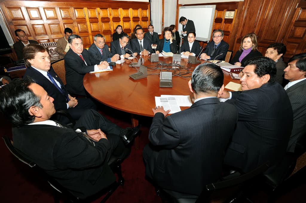 Eine Runde-Tisch-Besprechung mit Delegierten, die über Mikrofone, Papiere, Kabel und Dateien auf dem Tisch diskutieren, mit einer hölzernen Wand im Hintergrund.