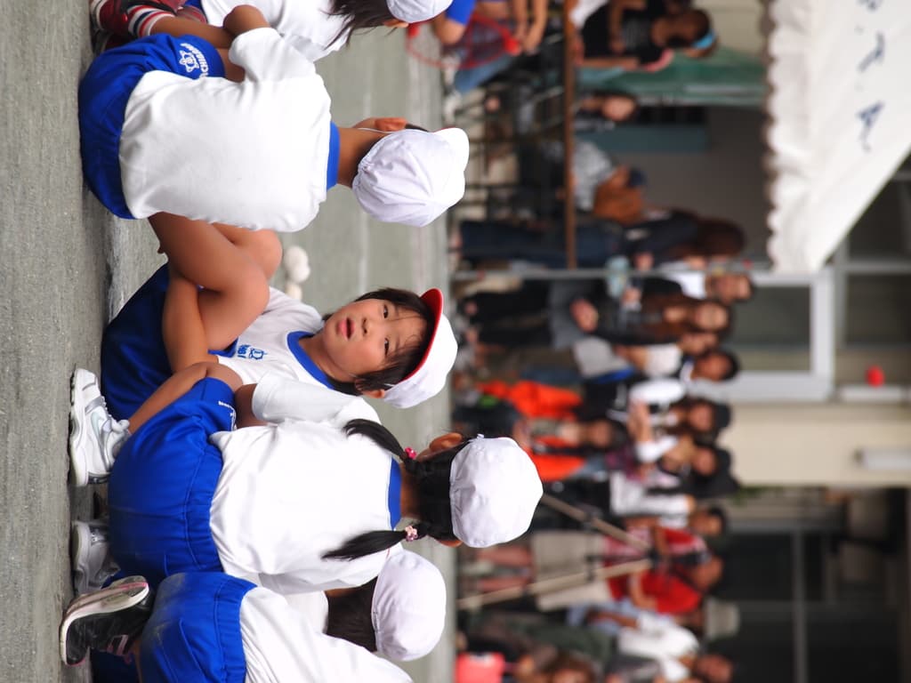 Kinder auf der Straße sitzend, im Hintergrund Menschen, ein Stativ, ein Zelt und eine Glaswand