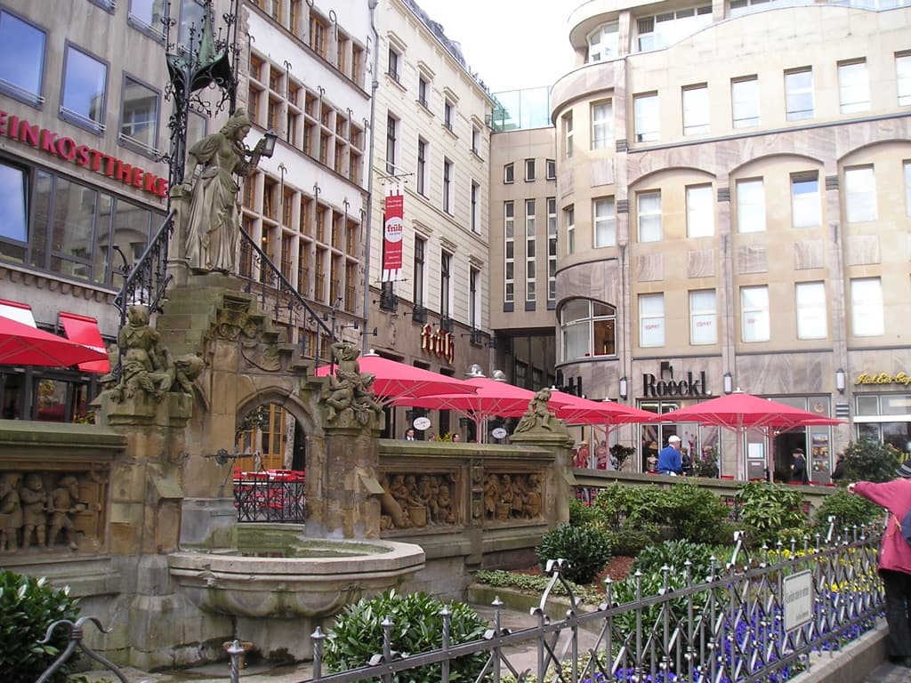 Eine Stadtlandschaft mit Gebäuden, Zelten, Pflanzen und Menschen im Hintergrund und einer Skulptur, einem Teich, einem Geländer und Pflanzen im Vordergrund.