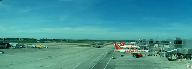 Eine Flughafen-Szene mit Flugzeugen, Gebäuden, Masten, Fahrzeugen und Bäumen unter einem Himmel mit einer Straße unten und einem Zaun im Vordergrund.