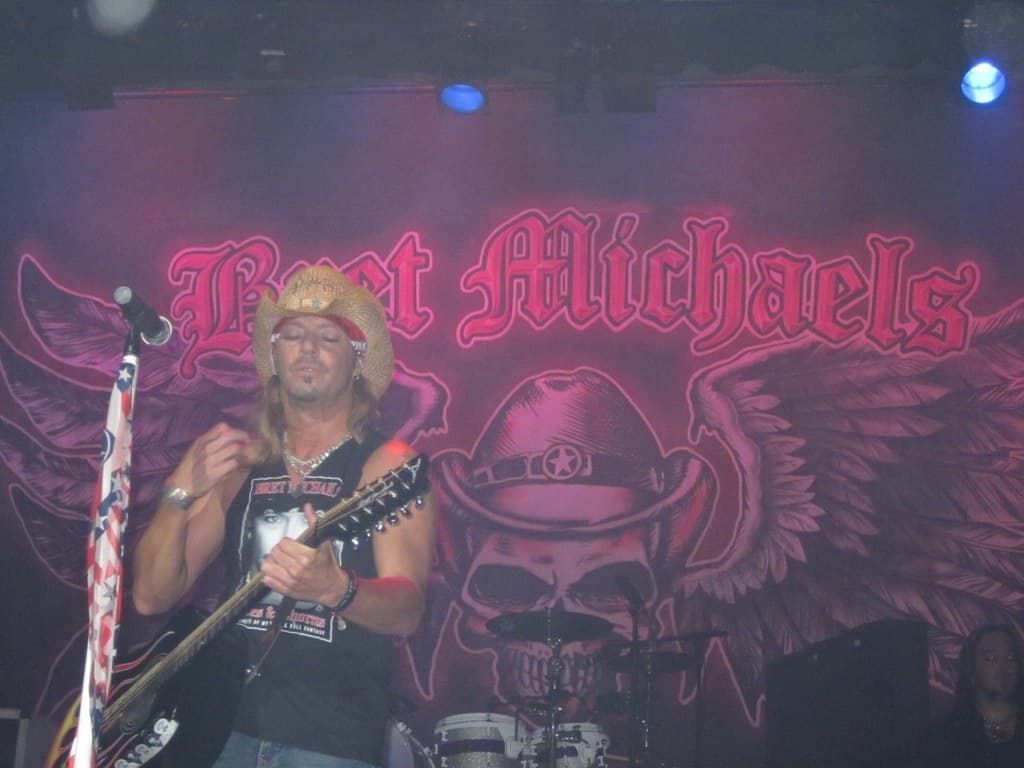 Eine Musikkonzert-Szene mit Scheinwerfern oben, einem Banner im Hintergrund, Drogen in der Mitte unten, einer Person in der rechten unteren Ecke und einem Gitarristen mit Mikrofon links.