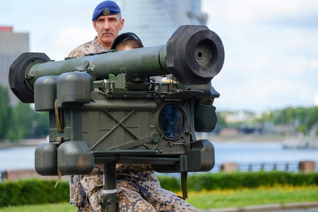 Zwei Personen in militärischer Uniform mit grünen Waffen, vor einem See, Gebäuden, Bäumen und einem klaren Himmel.