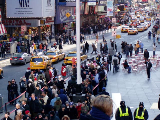 Fahrzeuge fahren auf einer Straße mit Fußgängern auf den Gehwegen, umgeben von Geschäften, Tischen, Stühlen, Laternen, Schildern und Ampeln.