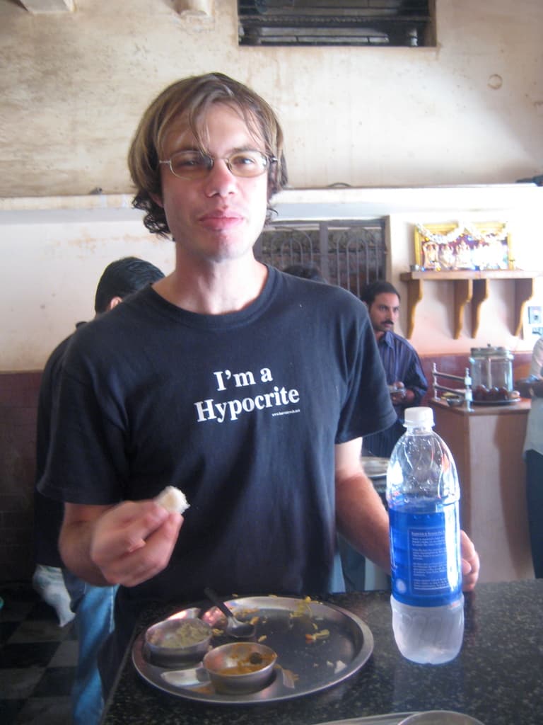 Eine Person sitzt an einem Tisch in einem Hotel, hält Essen in ihrer rechten Hand und hat eine Flasche auf dem Tisch, während im Hintergrund andere Menschen, ein Gott-Idol, ein Fenster und eine Wand zu sehen sind.