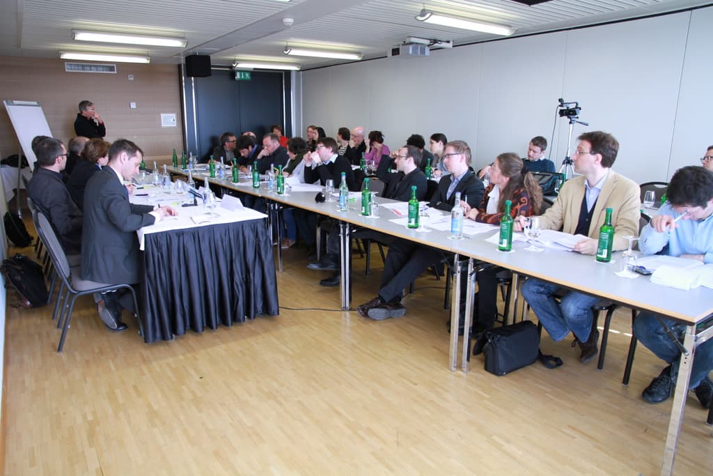 Ein Büroraum mit Angestellten an Tischen sitzend, Flaschen und Papieren auf den Tischen, Holzfußboden, Rucksäcke, ein stehender Mann im Hintergrund, eine Kamera, Lautsprecher, Schilder, Projektor, Deckenlampen, ein schwarzer Vorhang und viele Männer in Anzügen.