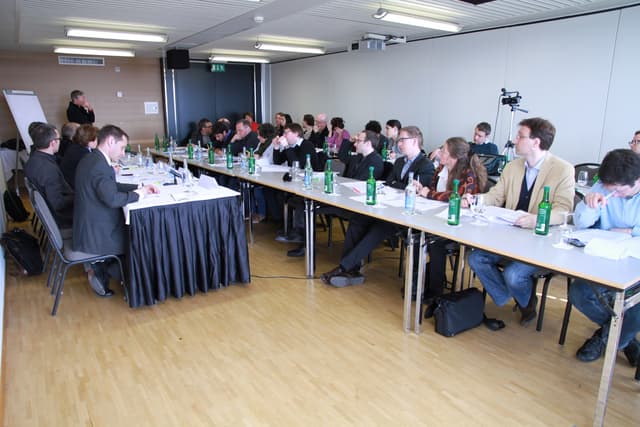 Ein Büroraum mit Angestellten an Tischen sitzend, Flaschen und Papieren auf den Tischen, Holzfußboden, Rucksäcke, ein stehender Mann im Hintergrund, eine Kamera, Lautsprecher, Schilder, Projektor, Deckenlampen, ein schwarzer Vorhang und viele Männer in Anzügen.