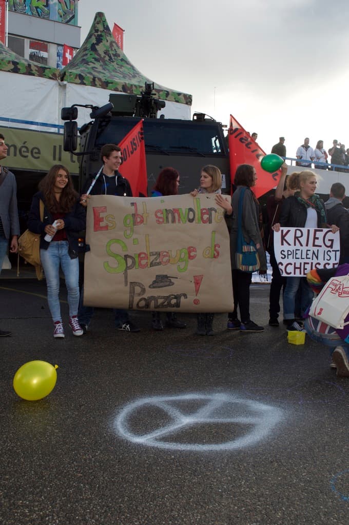 Eine Stadtstraßenszene mit einem Ballon im Vordergrund, Menschen mit Schildern und Fahnen in der Mitte, einem Gebäude und einem Zelt im Hintergrund, unter einem bewölkten Himmel.
