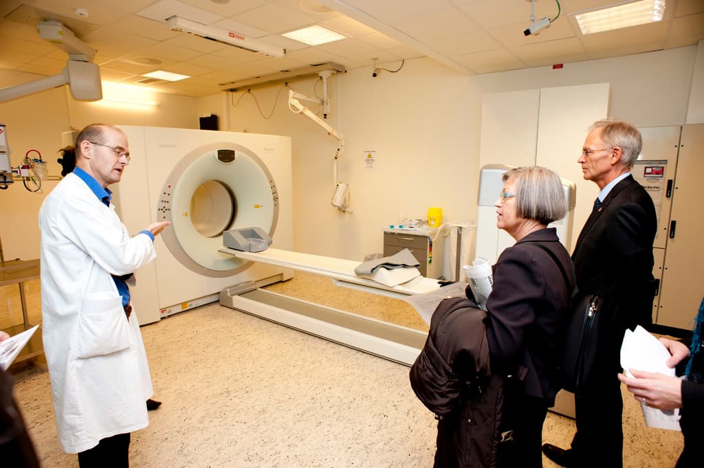 Ein Arzt in weißer Uniform erklärt einem CT-Scan-Gerät zwei Personen in schwarzen Mänteln.
