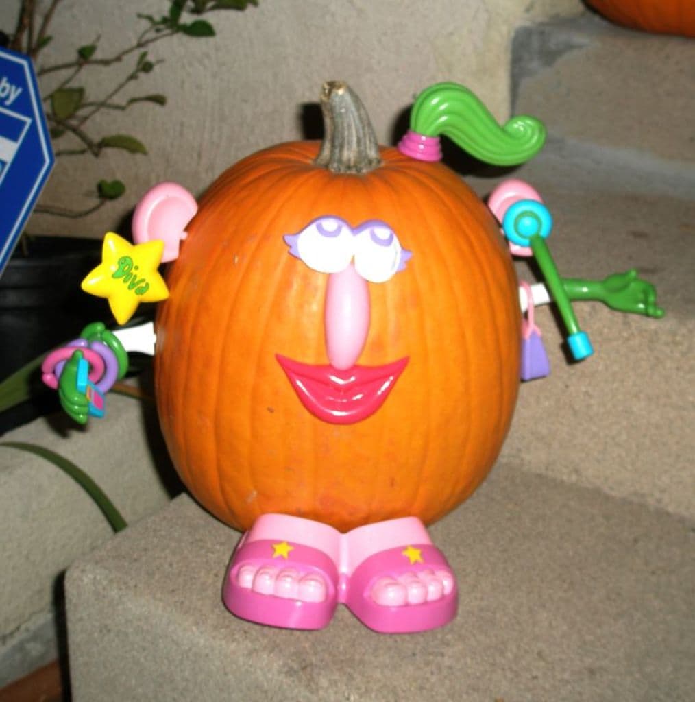 Ein Kürbis steht auf einer Treppe mit Spielzeugen oben drauf, eine Pflanze und ein blaues Objekt links daneben und ein weiterer Kürbis darüber.