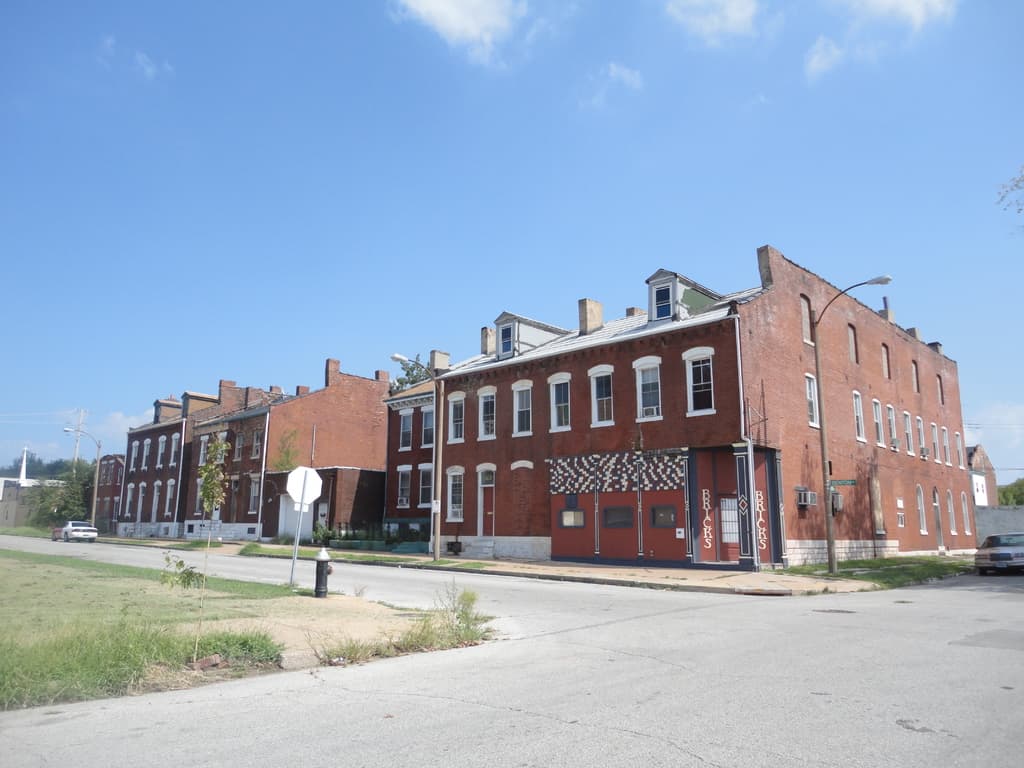 Ein Gebäude mit Masten und Straßenlaternen, umgeben von Bäumen und Fahrzeugen auf beiden Seiten, mit einem Weg, Gras und einem Feuerlöscher unten und dem Himmel oben.