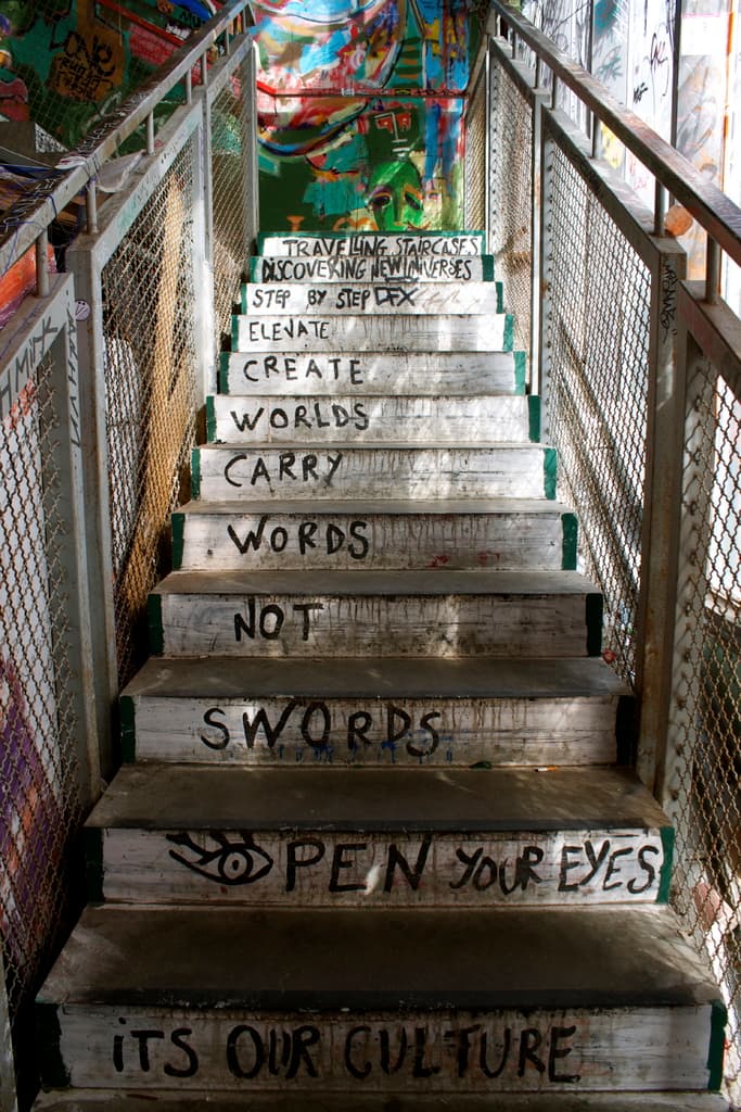 Eine Treppe mit Text auf den Stufen, flankiert von Metallgeländern auf beiden Seiten, mit einer Wand und einem Gemälde im Hintergrund.