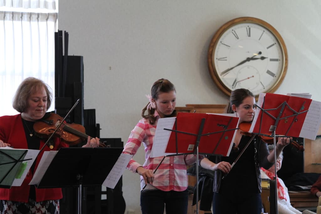 Drei Frauen auf der Bühne mit Geigen stehend und Notenständer vor sich, während ein sitzendes Publikum zuhört; eine Uhr ist an der weißen Wand im Hintergrund angebracht, und ein weißer Vorhang ist links sichtbar.