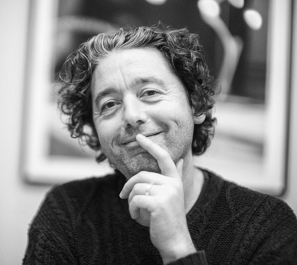 Ein Mann in einem Sweatshirt l├Ąchelt, mit einem gerahmten Bild an der Wand dahinter.