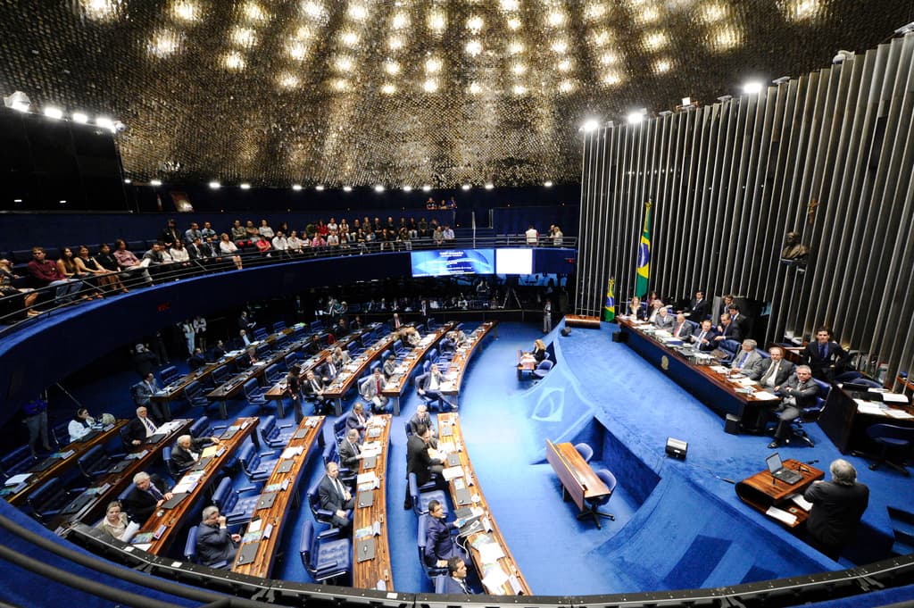 Eine Konferenzbühne mit sitzenden Personen an einem Tisch mit Papieren und Gegenständen, flankiert von Flaggen und einer Skulptur im Hintergrund, mit Zuschauern an Tischen und hinter einer Absperrung mit Bildschirmen, unter dekorativen Deckenleuchten.