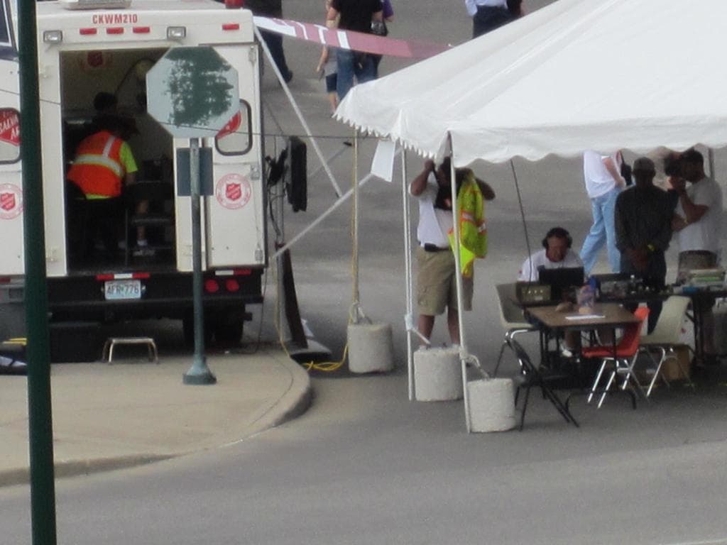 Ein Krankenwagen steht auf der linken Seite einer Straße, während auf der rechten Seite ein Zelt viele Menschen schützt, mit einem Tisch und einem Stuhl in der Nähe.