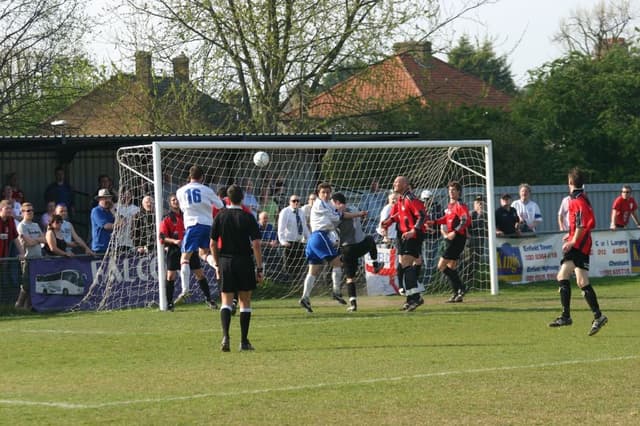 Fußballspieler spielen auf einem Feld mit einem Tor, während Zuschauer dahinter stehen, mit Bäumen und Häusern im Hintergrund.