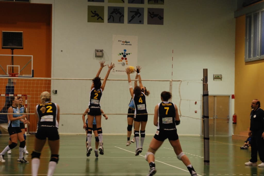 Ein Volleyballspiel in vollem Gange auf einem Indoor-Court, mit aktiv beteiligten Spielern, einem Basketballkorb im Hintergrund und verschiedenen architektonischen Elementen wie Rahmen, Wänden, Fenstern und einer Tür.