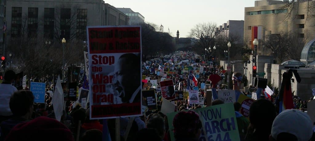 Eine Gruppe von Menschen, die auf einer Straße protestieren und Schilder halten, mit Gebäuden, Bäumen, Laternenmasten und dem Himmel im Hintergrund.
