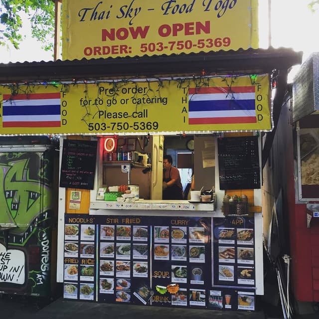 Ein Geschäft mit verschiedenen Artikeln wie Getränkedosen, Speisekarten, einer Uhr, Gewürzen in Plastikbehältern, Gewürzen, Werbetafeln und Namensschildern unter einem Himmel.