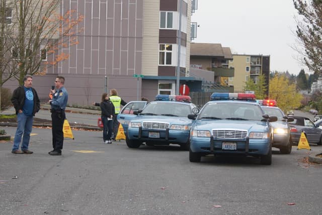 Autos auf einer Straße mit vier Personen in der Nähe, Gebäude mit Fenstern im Hintergrund, Bäume und Warnwesten.