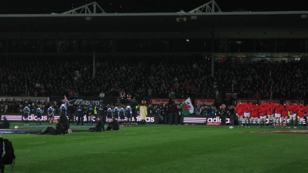 Eisernes Gitter und Lautsprecher oben auf dem Bild, mit Menschenmenge, Werbung, Flagge und Boden unten.