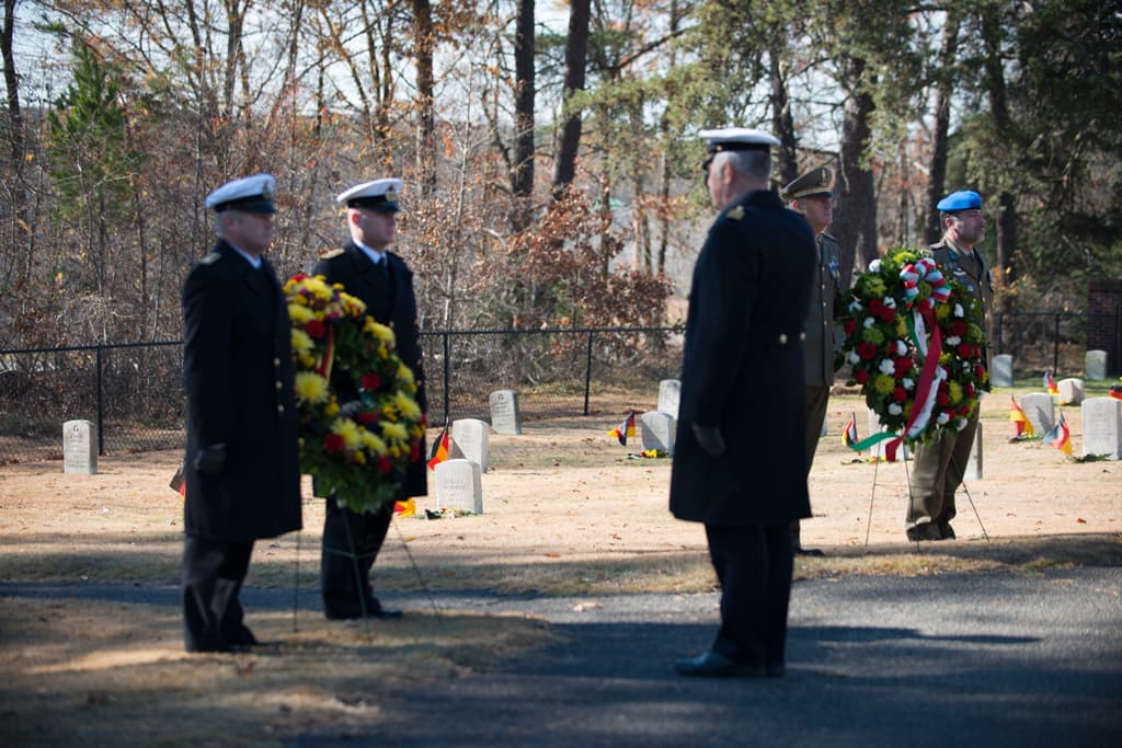 Zwei Beamte halten einen Blumenkranz und stehen auf einem Boden mit Flaggen und Steinen, während ein weiterer Beamter vor ihnen steht und zwei weitere Beamte einen Blumenkranz auf der rechten Seite halten, mit Bäumen im Hintergrund.