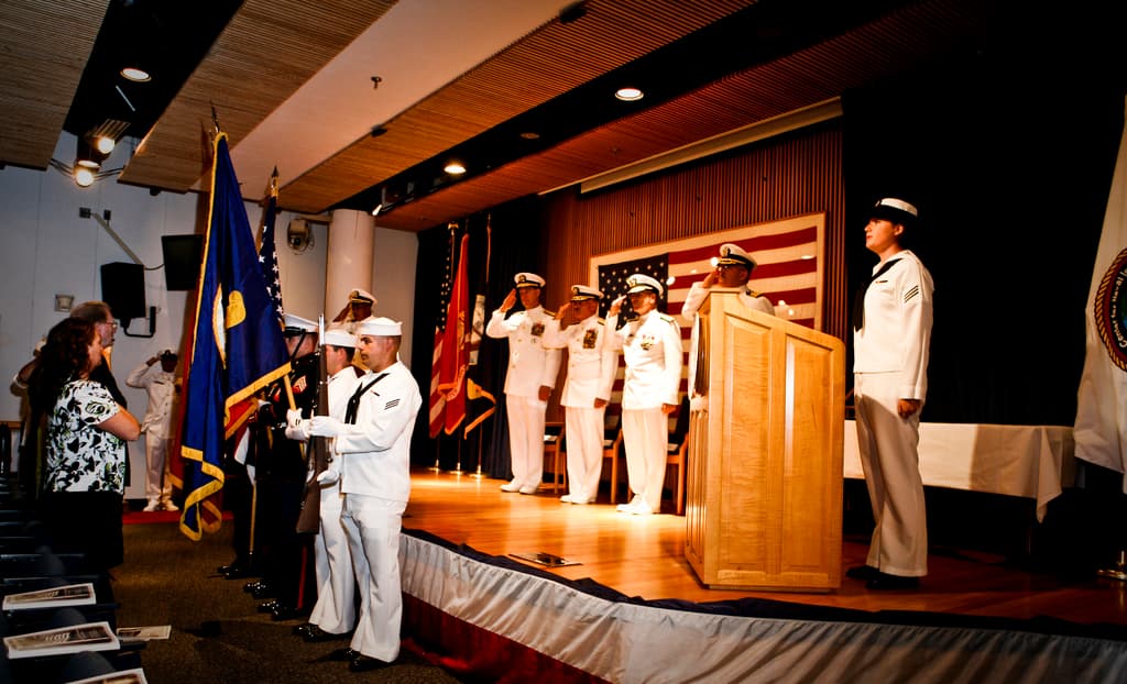 Eine formelle Veranstaltung mit Menschen, die auf einer Bühne stehen und salutieren, Flaggen in Rot, Blau und Weiß, Stühle, ein Podium und ein Tisch mit einer weißen Decke, mit einem Fernseher und einem Lautsprecherbox im Hintergrund.