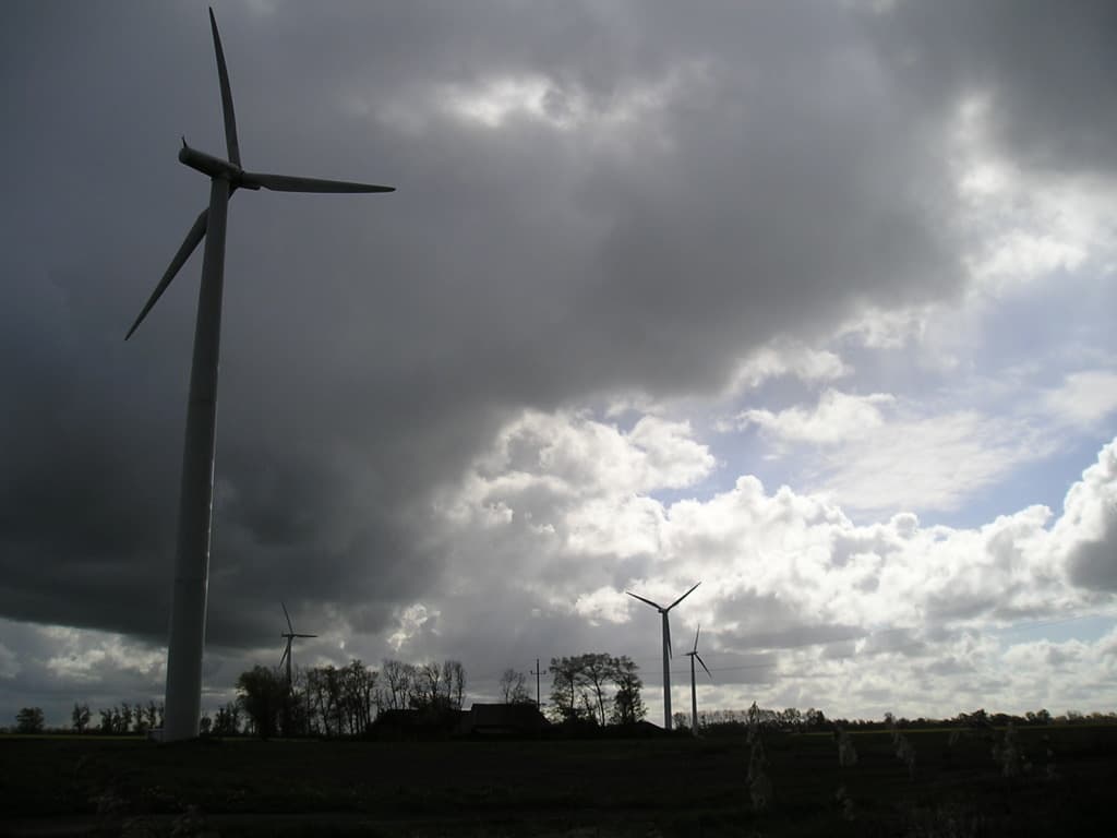 Windmühlen in einer Landschaft mit Bäumen, Wolken am Himmel und einem Haus in der Ferne.