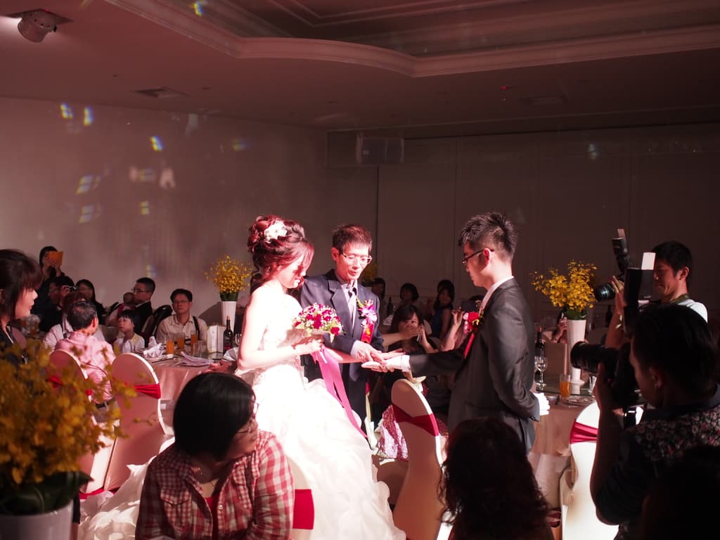 Ein Brautpaar steht in der Mitte eines Raumes mit Menschen, die Kameras halten, rechts, einige sitzen und einige stehen, mit Wänden, Lampen und Blumenvasen im Hintergrund.