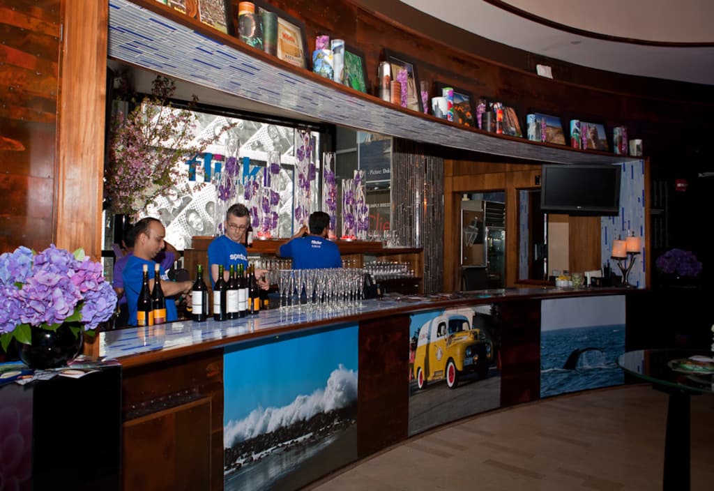 Ein Restaurantinnenraum mit drei Personen hinter einem Tisch, der mit Gläsern, Flaschen, Aufklebern, Postern, einer Zimmerpflanze und Lampen bedeckt ist, mit einem Fernseher und Regalen im Hintergrund.