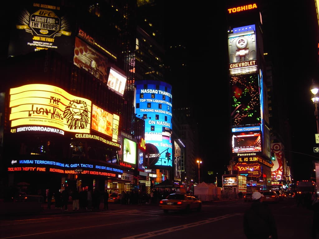 Eine Straßenszene bei Nacht mit fahrenden Fahrzeugen, Gebäuden mit beleuchteten Schildern und Straßenlaternen.