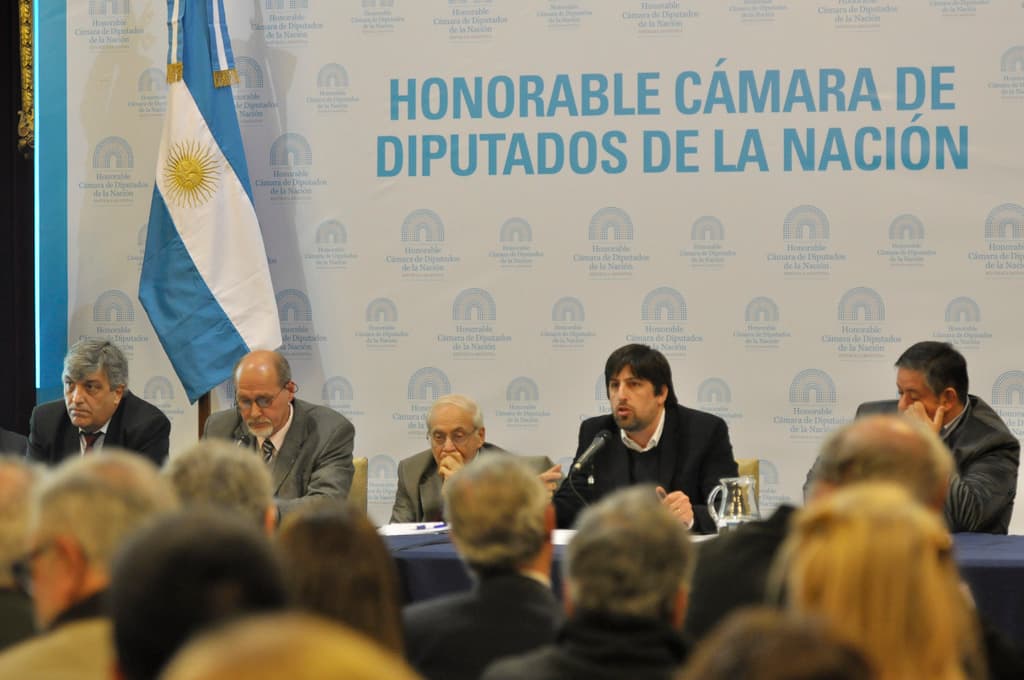 Eine Gruppe von Menschen an Tischen mit einem Krug, Papieren und einem Stift, während eine Person in der Mitte in ein Mikrofon spricht, mit einer weißen und blauen Flagge und einem Hintergrund, auf dem "ehrenwerte kamara de" steht.