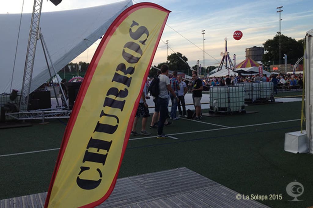 Eine lebhafte Karnevalszene mit Zelten, einem zentralen Mast, Menschen, Bäumen, Strommasten, Straßenbahnen auf der Straße und einem Eisenständer auf der linken Seite unter einem klaren Himmel.