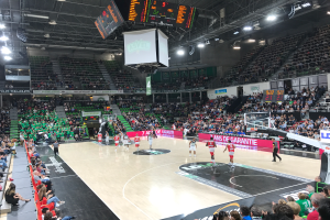 Ein Basketballspiel in einer großen Arena mit Zuschauern auf den Tribünen, Anzeigetafeln mit "Asvel vs Galatasaray" und einer beleuchteten Decke mit einem Bildschirm über dem Spielfeld.