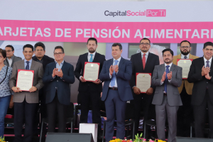 Gruppe von Menschen auf einer Bühne, die klatschen und Zertifikate halten, mit Stühlen in einem Halbkreis angeordnet, ein Banner mit der Aufschrift "Capital Social for Ti" im Hintergrund und Blumensträuße und Lautsprecher unten.