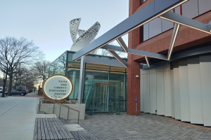 Eingang zum Faith and Liberty Discovery Center mit Glaswänden und -türen, einem Namensschild, einer Bank, einem Metallrahmen, Bäumen, parkenden Fahrzeugen, Pfosten und einem bewölkten Himmel.